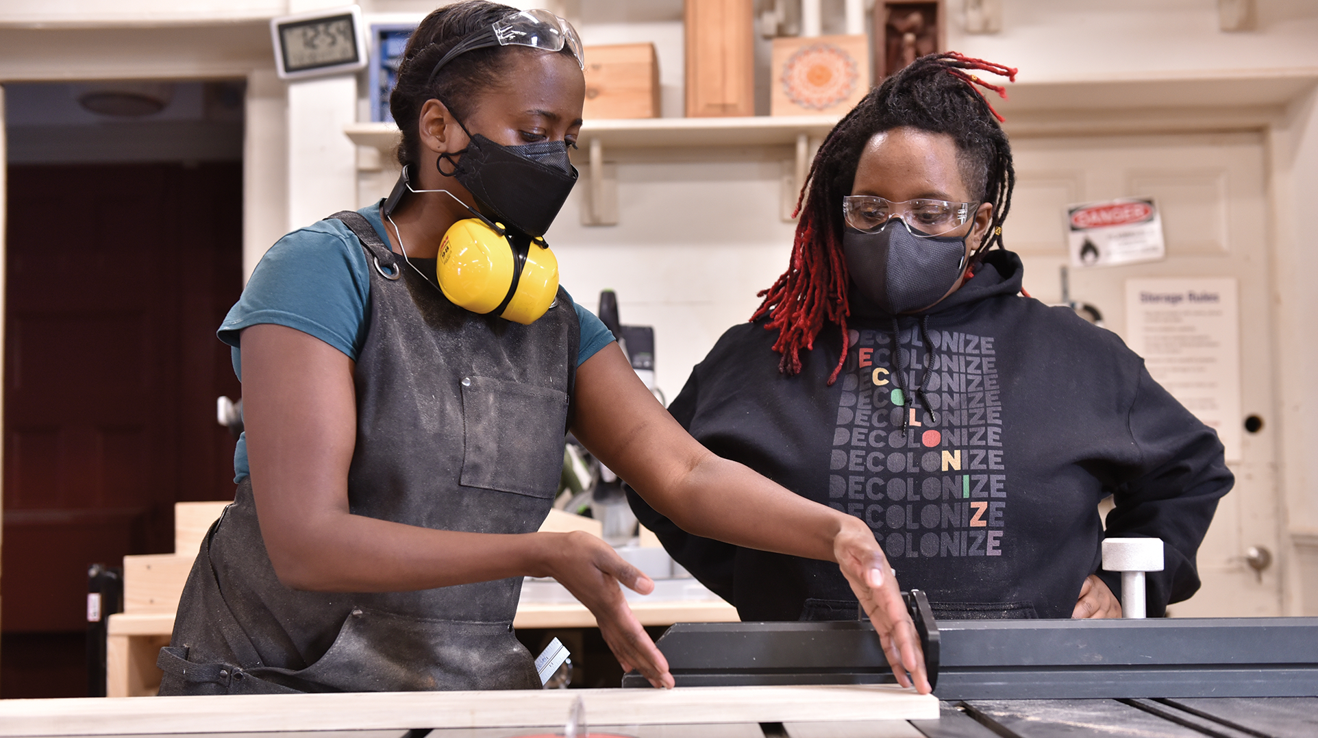 Two masked people collaborate on a woodworking project, one wearing a sweatshirt that says 