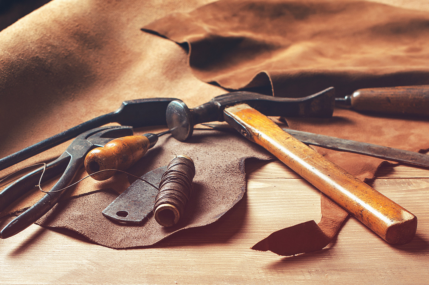 Leather working tools including a hammer, pliers, awl, and cordage laying atop a pile of leather.