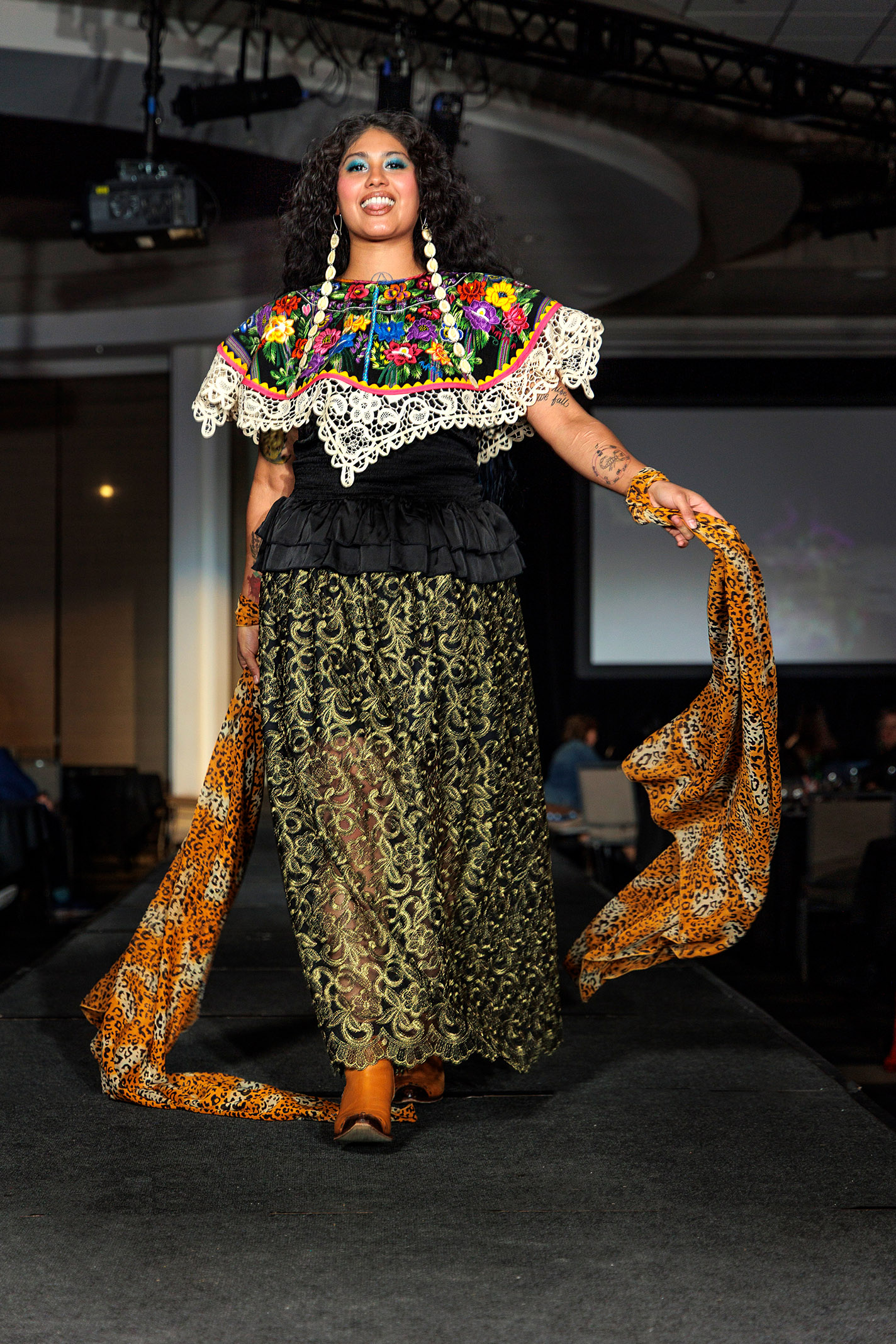Model wears translucent skirt, floral cape, and long earrings on the runway.