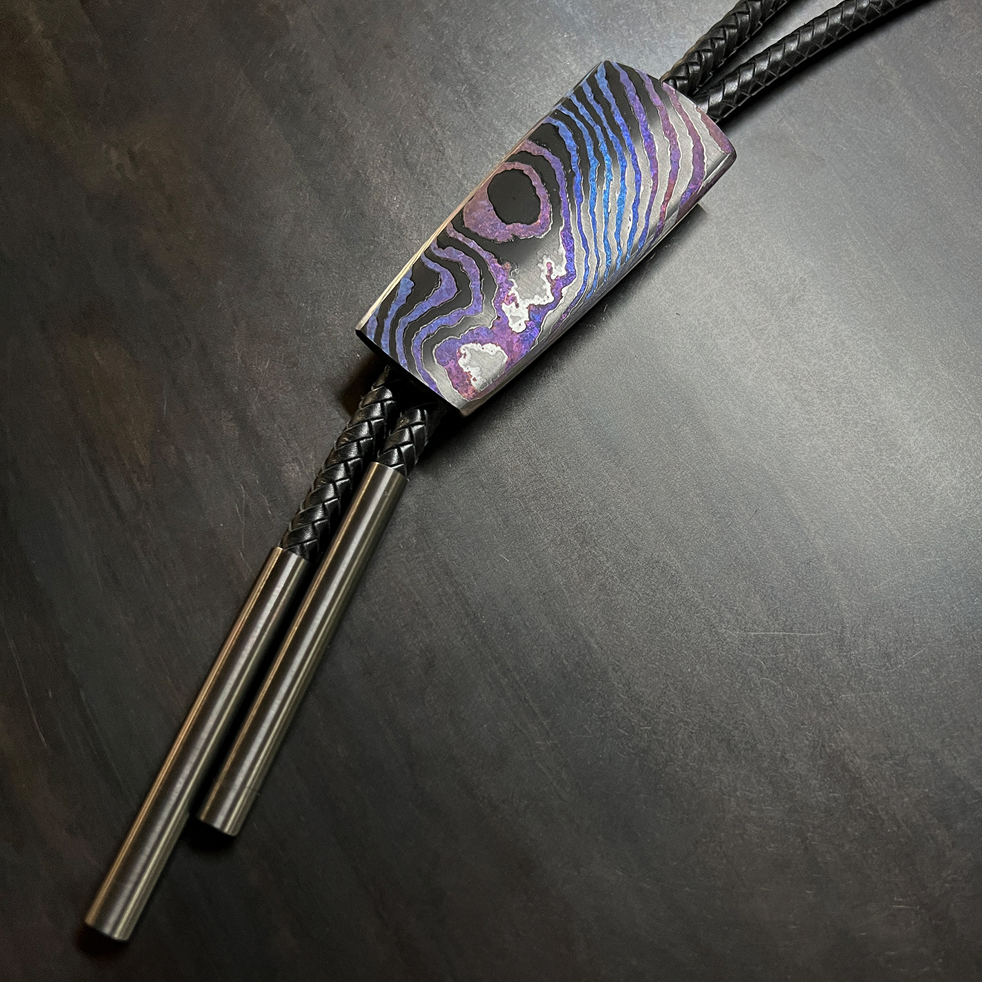 Bolo pendant made with a custom laminate of zirconium and titanium with iridescent patterns on a braided kangaroo-leather cord.